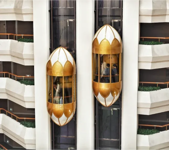 Two modern gold and white capsule elevators ascending within a multi-story building atrium, with balconies and green foliage visible on the surrounding floors