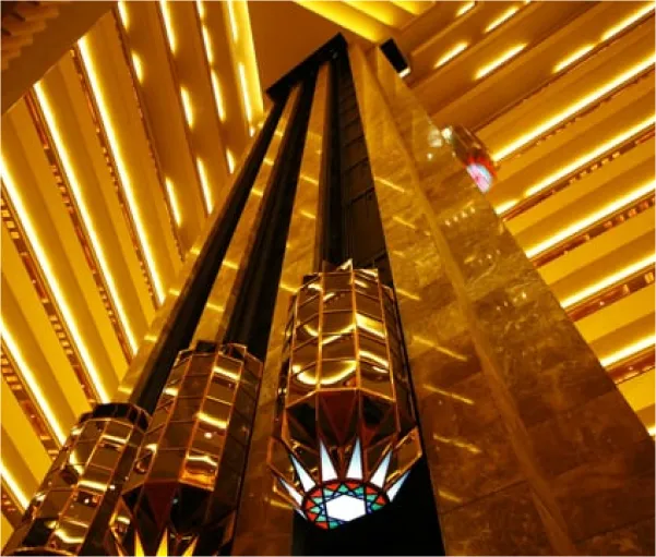 Looking up at multiple hydraulic glass elevators ascending within a luxurious, gold-toned interior of a modern building, possibly a hotel or shopping center, featuring a striking architectural design.